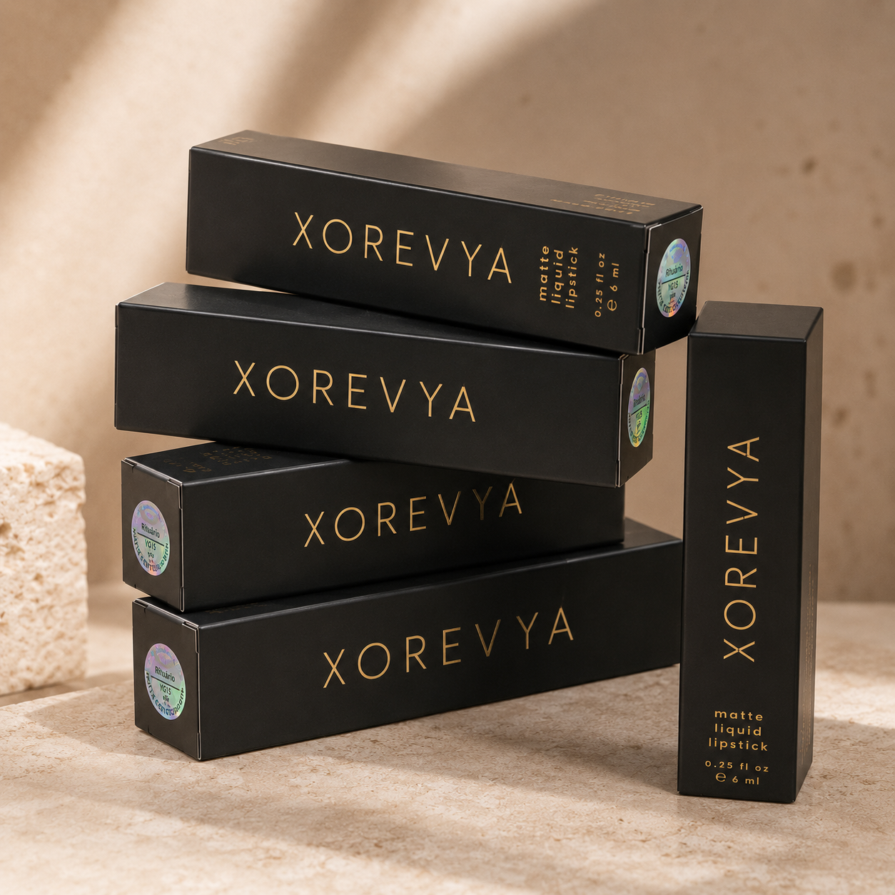 Four black XOREVYA cosmetic boxes stacked and arranged on a beige stone surface with soft warm lighting. The packaging is matte black with gold lettering and small circular green-and-white stickers on the ends.