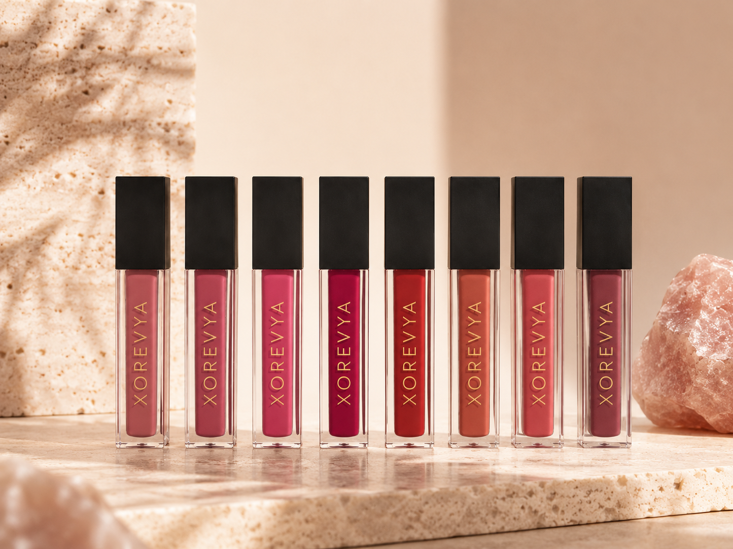 Seven XOREVYA liquid lipstick tubes in various shades of pink, red, and nude, lined up on a beige stone surface with a warm beige background and decorative rocks.