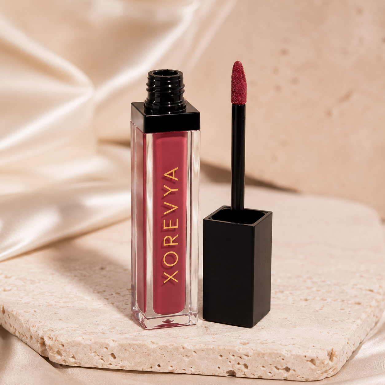 Xorevya liquid lipstick in a rectangular clear bottle with a black cap, shown next to the applicator wand on a beige stone slab with draped satin fabric in the background.