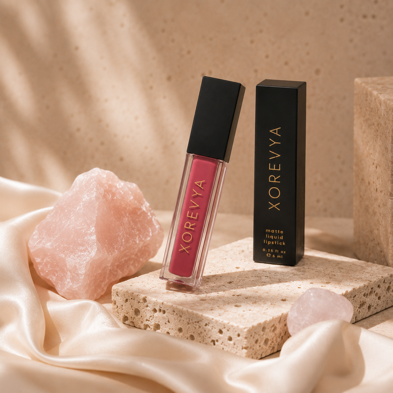 Xorevya matte liquid lipstick in a pink shade, shown in a clear tube with a black cap next to its black packaging box, styled on a stone pedestal with pink crystal rocks and draped satin fabric.